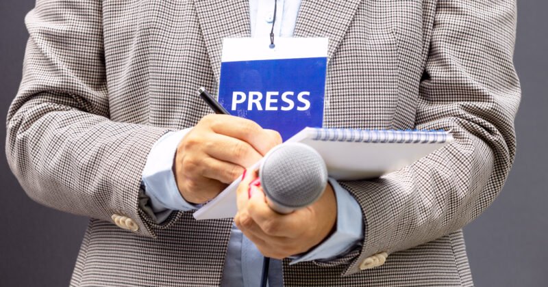 Female journalist at news conference or media event, writing notes, holding microphone